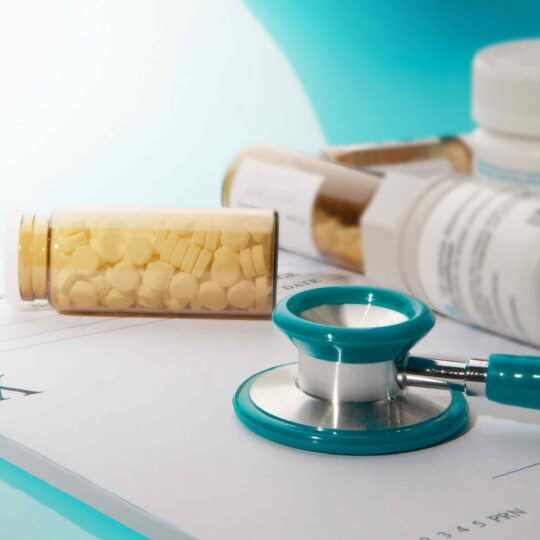 Un frasco de pastillas blancas con tapa azul y un estetoscopio negro sobre una receta médica escrita en papel blanco. La imagen representa la atención médica, la enfermedad y la medicación.